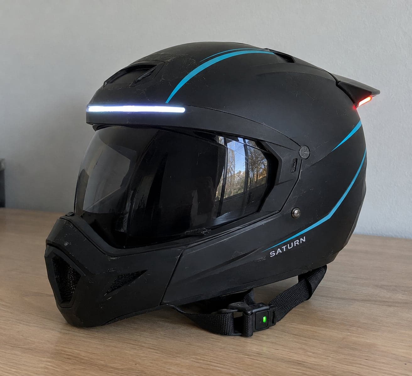 Concept image of the Saturn Smart Kids' E-Bike Helmet on a wooden table: a matte black full-face helmet with thin cyan stripes, a white light across the brow, a red light on the rear spoiler, and a green light on the chin strap.